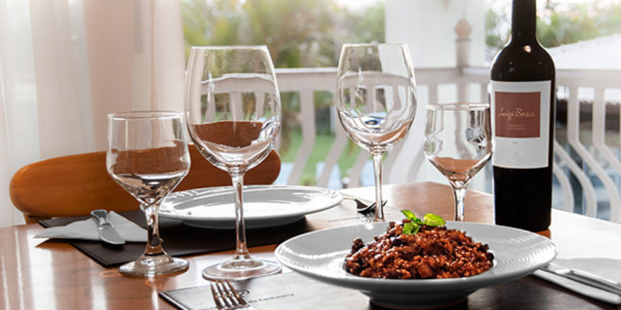 mesa posta com dois pratos, sendo um servido com risoto, quatro taças, talheres e uma garrafa de vinho.