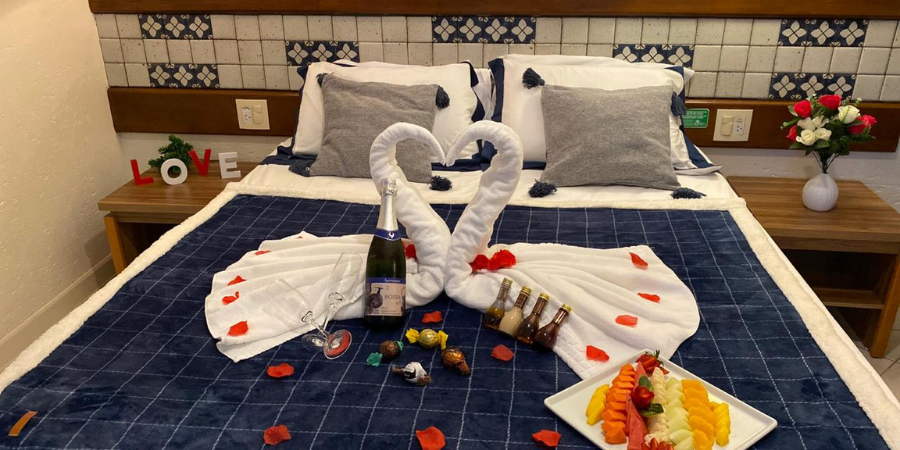 decoração romântica com toalhas em forma de cisnes, prato de frutas, óleos, bombons e bebida em cima da cama.