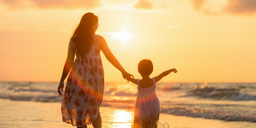 mãe e filha caminhando na beira da praia de mãos dadas ao pôr do sol