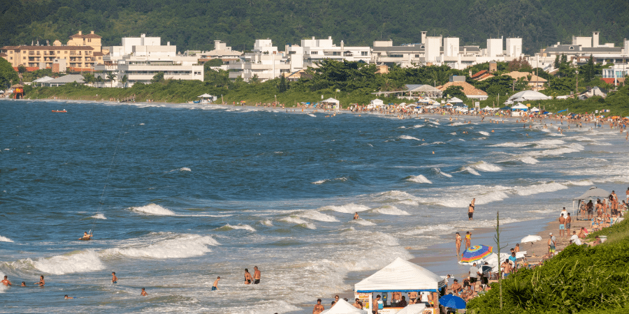 Diversas pessoas na beira da praia, mar com ondas e prédios baixos ao fundo
